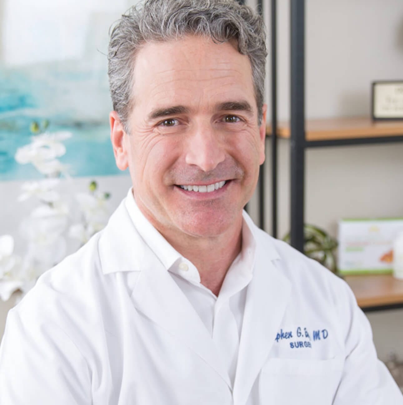 Smiling male surgeon in a white coat, seated in an office with plants and diplomas in the background.